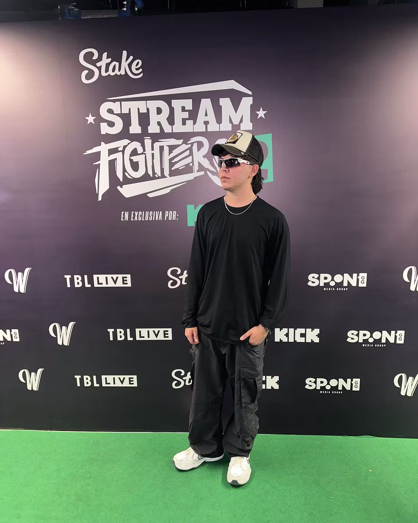 Trianiss en Stream Fighters, con gorra, gafas oscuras y ropa negra frente al muro de prensa del evento